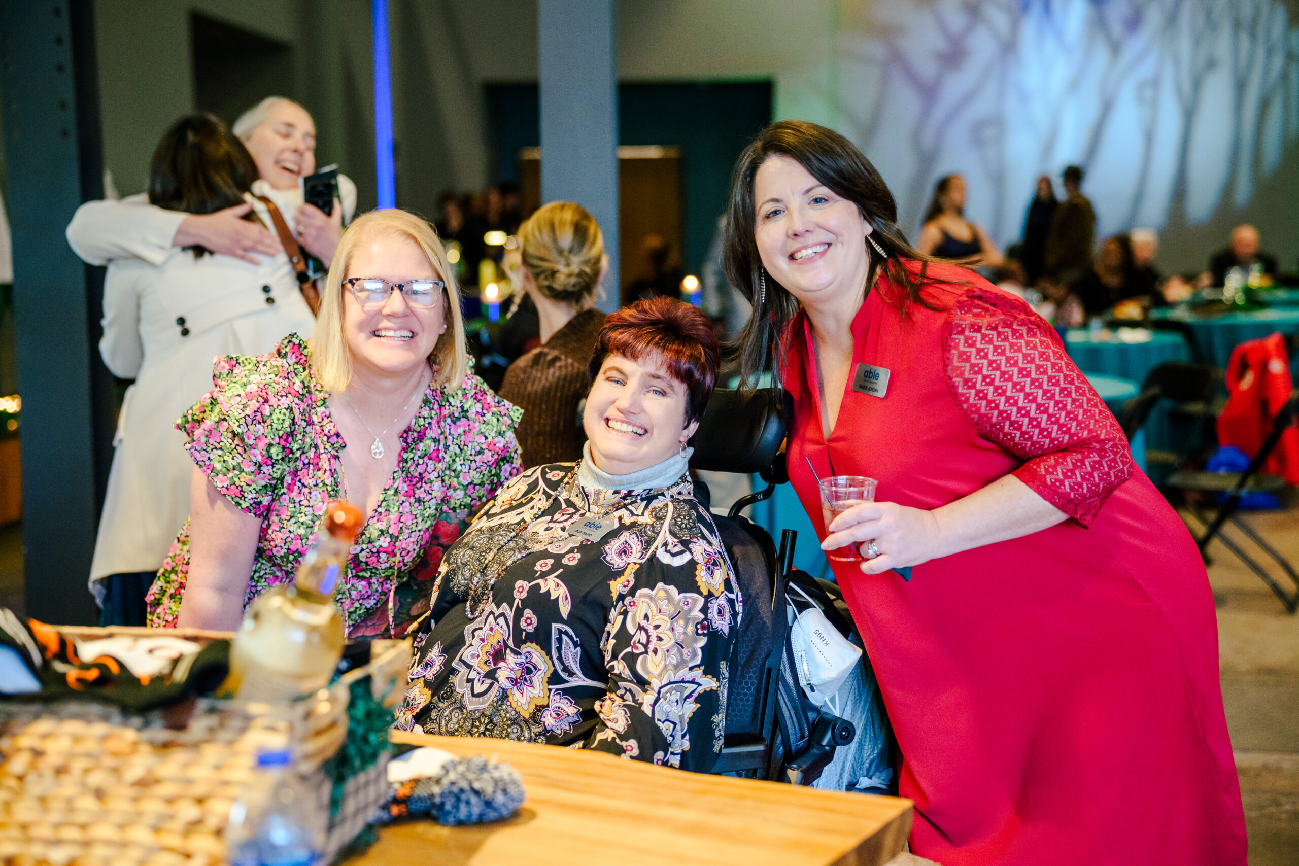 Keri, a white person in a floral dress and glasses, Dori, a white woman using a power wheelchair, and Sandy, a white woman in a red dress, pose and smile together for a photo.